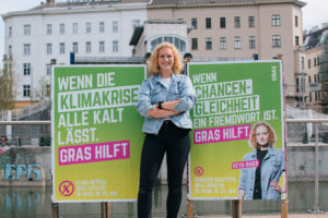 GRAS-Spitzenkandidatin Keya Baier mit zwei Plakaten. Plakat 1: "Wenn die Klimakrise alle kalt lässt. GRAS hilft." Plakat 2: "Wenn Chancengleichheit ein Fremdwort ist. GRAS hilft"